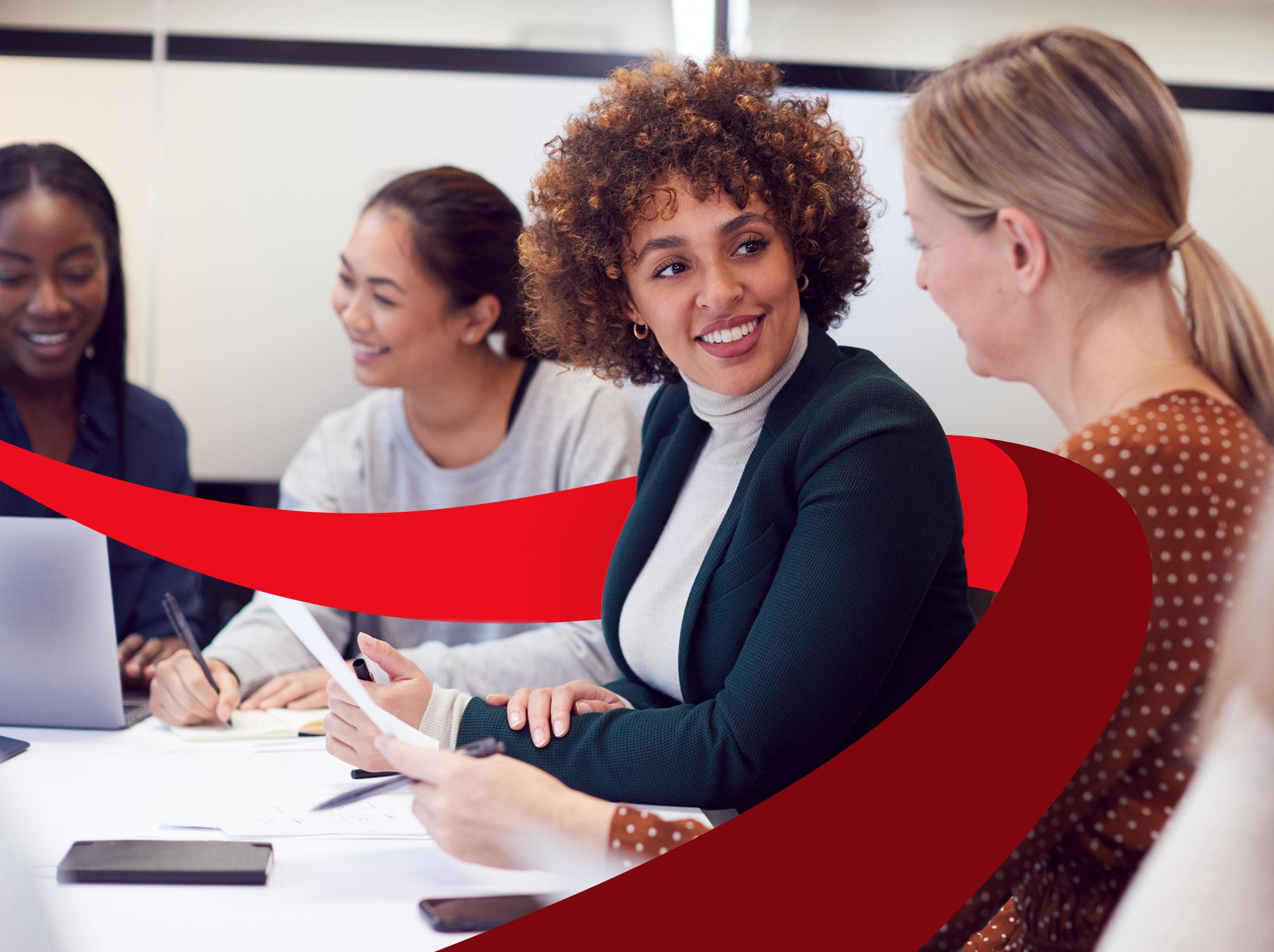 Group of women leaders talking amongst themselves around a conference table with laptops and papers. They are smiling and having pleasant conversation.