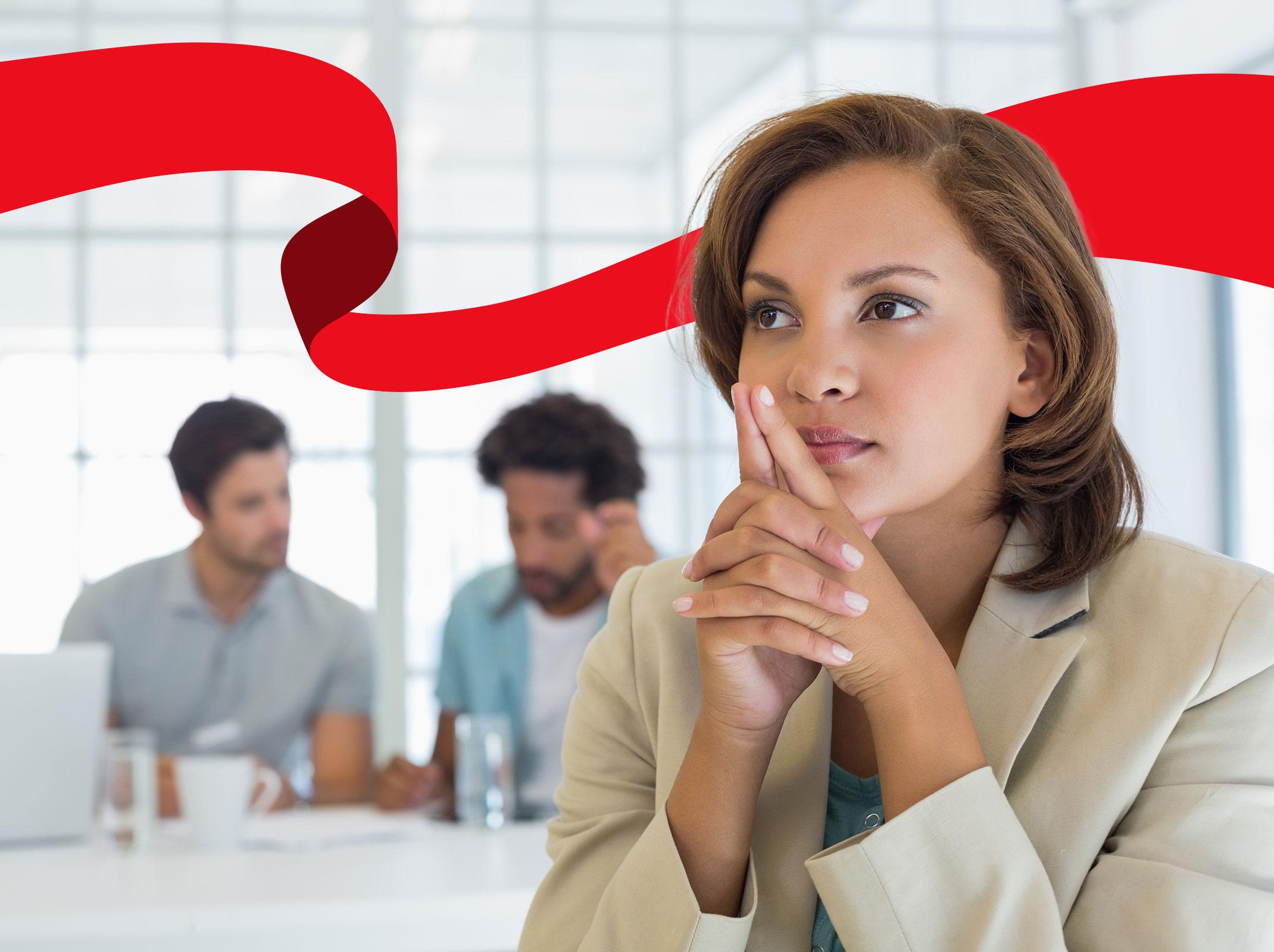 A business professional with her fingers crossed near her face.  There are other business professionals in the background.  She appears to be thinking.